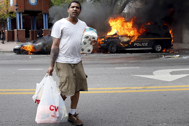 A man with goods looted from a store walks past burning vehicles during clashes in Baltimore, Maryland